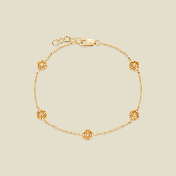 A delicate gold chain bracelet featuring five ornate, textured gold poppy charms with small clear stones, arranged in a circle on a light beige background.