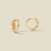 A pair of gold hoop earrings adorned with textured poppy flowers stacked along the hoop, with scattered small clear accent stones, set against a solid beige background.