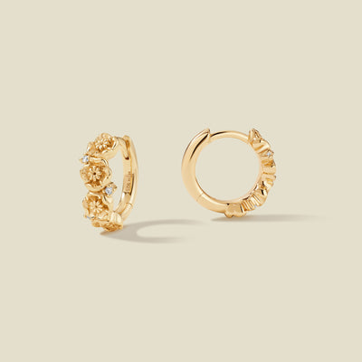 A pair of gold hoop earrings adorned with textured poppy flowers stacked along the hoop, with scattered small clear accent stones, set against a solid beige background.