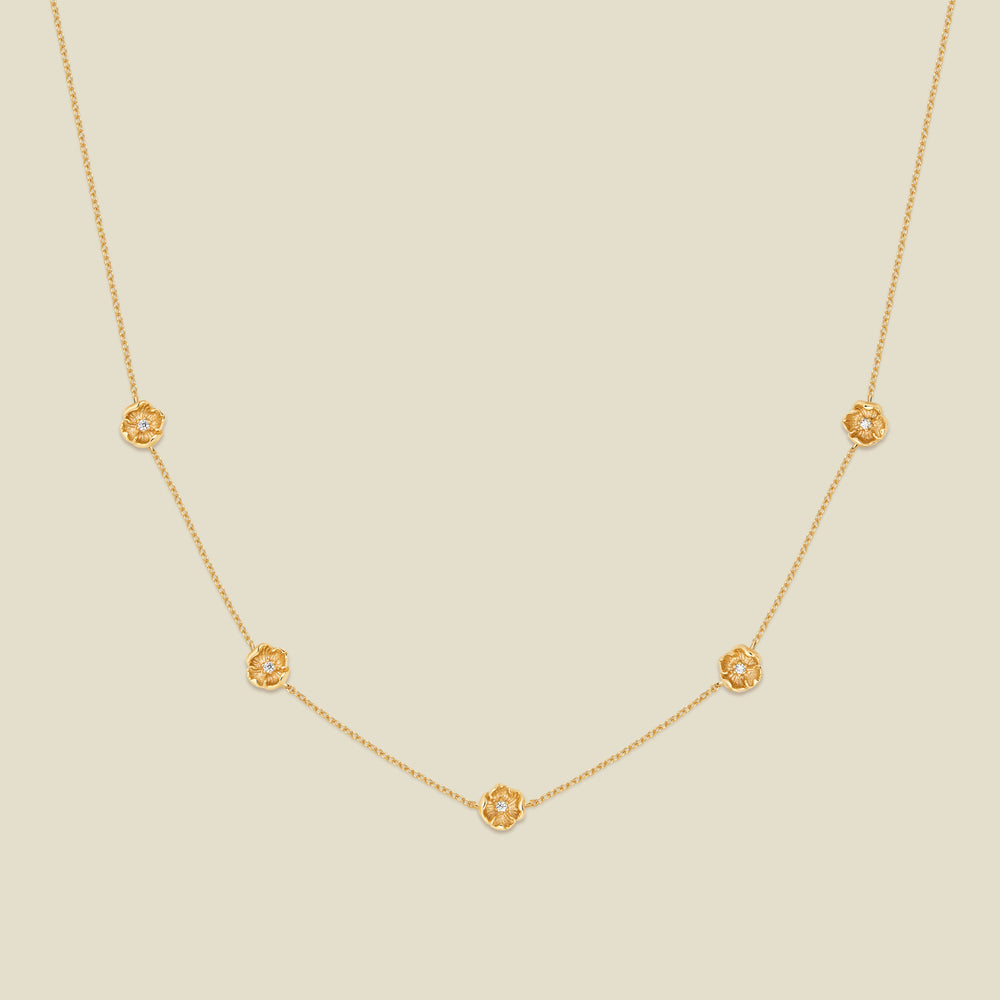 A delicate gold chain necklace featuring five small, evenly spaced textured poppy flower motifs set with tiny clear stones, displayed on a solid light beige background.