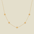 A delicate gold chain necklace featuring five small, evenly spaced textured poppy flower motifs set with tiny clear stones, displayed on a solid light beige background.