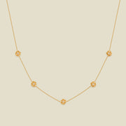 A delicate gold chain necklace featuring five small, evenly spaced textured poppy flower motifs set with tiny clear stones, displayed on a solid light beige background.