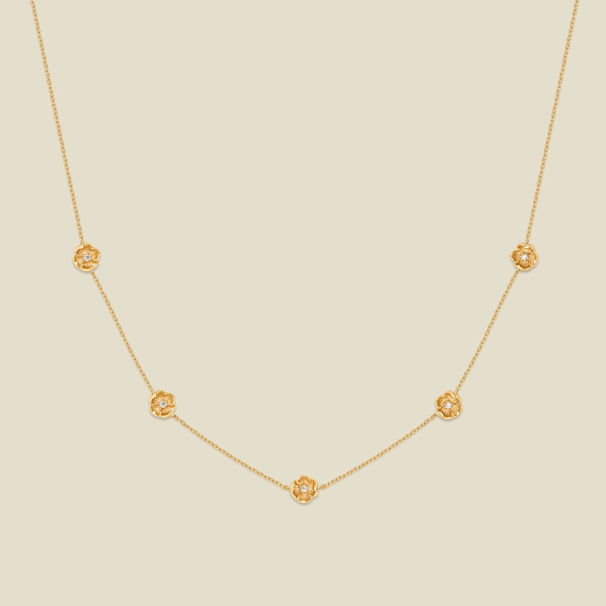 A delicate gold chain necklace featuring five small, evenly spaced textured poppy flower motifs set with tiny clear stones, displayed on a solid light beige background.