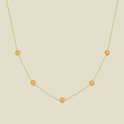 A delicate gold chain necklace featuring five small, evenly spaced textured poppy flower motifs set with tiny clear stones, displayed on a solid light beige background.