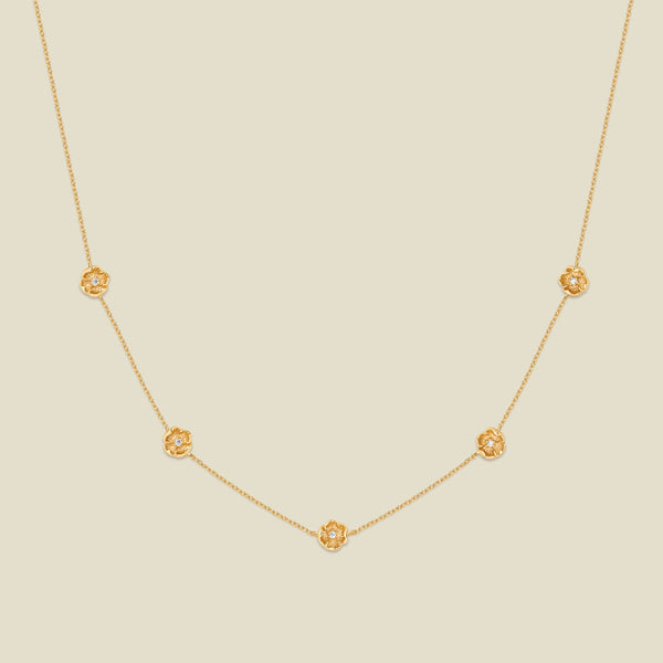 A delicate gold chain necklace featuring five small, evenly spaced textured poppy flower motifs set with tiny clear stones, displayed on a solid light beige background.