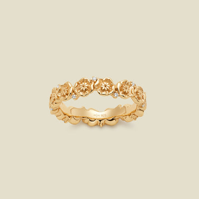 A gold floral-etched ring band with textured poppy flowers and small clear gemstones, shown on a plain warm beige background. 