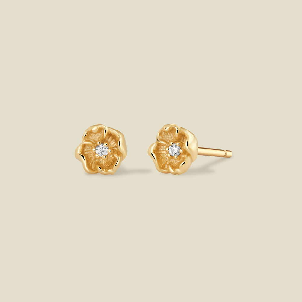 A pair of gold poppy flower stud earrings, each featuring a small round clear gemstone at the center, displayed on a solid light beige background.