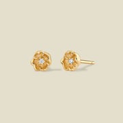 A pair of gold poppy flower stud earrings, each featuring a small round clear gemstone at the center, displayed on a solid light beige background.