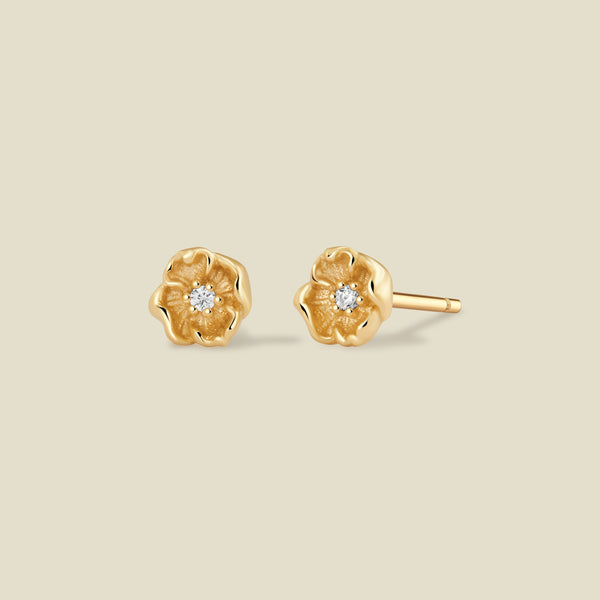 A pair of gold poppy flower stud earrings, each featuring a small round clear gemstone at the center, displayed on a solid light beige background.