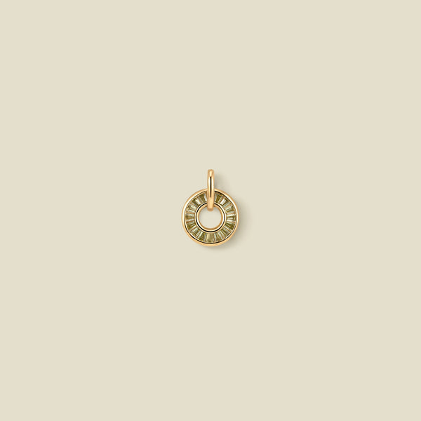 A circular gold pendant featuring light green baguette-cut stones around a hollow center, displayed against a solid light beige background.