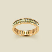 A gold eternity ring featuring a continuous channel setting of baguette-cut light green gemstones, shown on a solid light beige background.