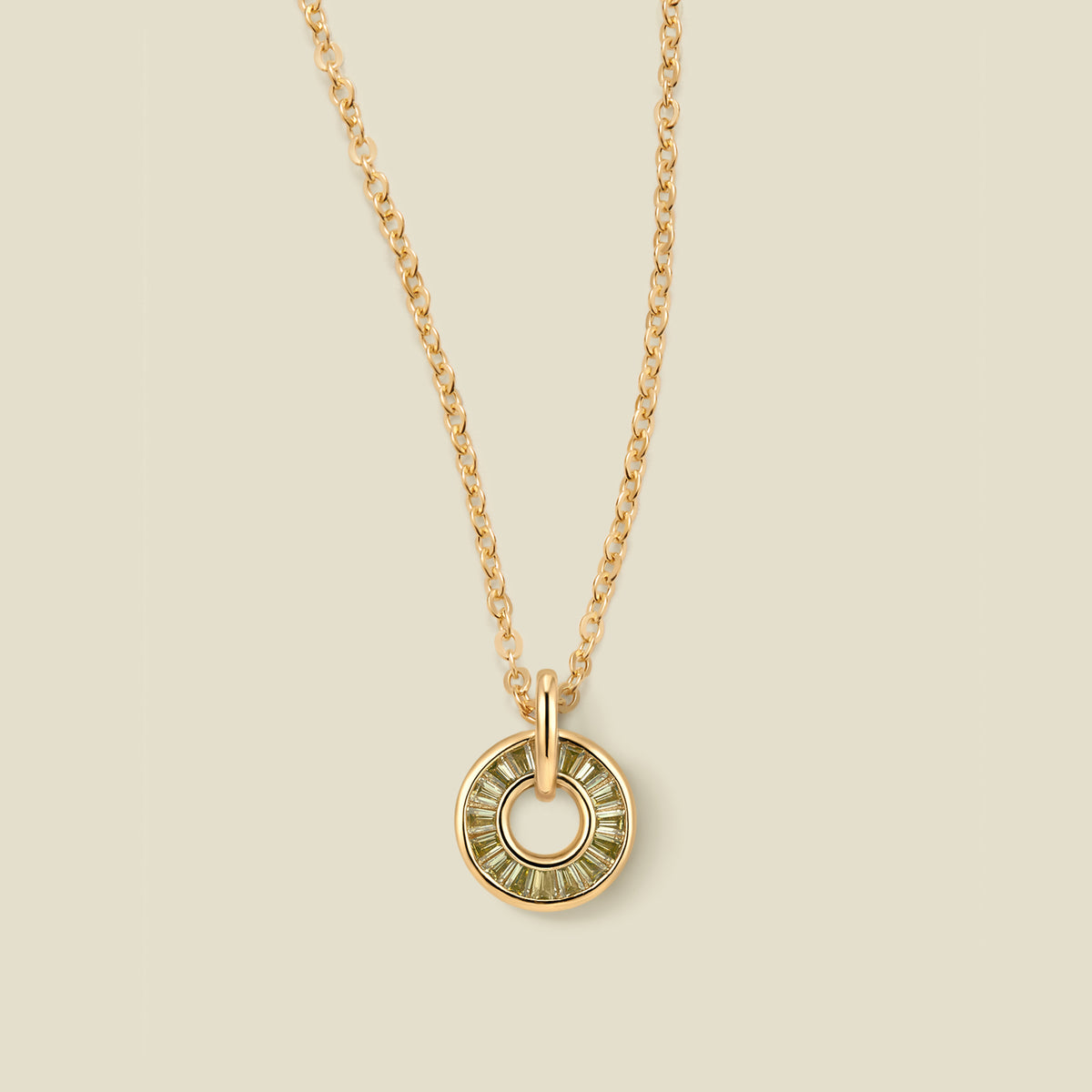 A gold chain necklace with a circular pendant featuring light green baguette-cut stones around a hollow center, displayed against a solid light beige background.