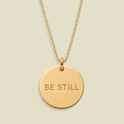 A gold disc pendant necklace engraved with the words 'BE STILL' in a clean sans-serif font, suspended from a delicate gold chain against a solid, warm beige background.