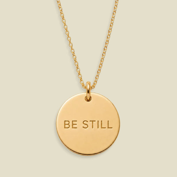 A gold disc pendant necklace engraved with the words 'BE STILL' in a clean sans-serif font, suspended from a delicate gold chain against a solid, warm beige background.