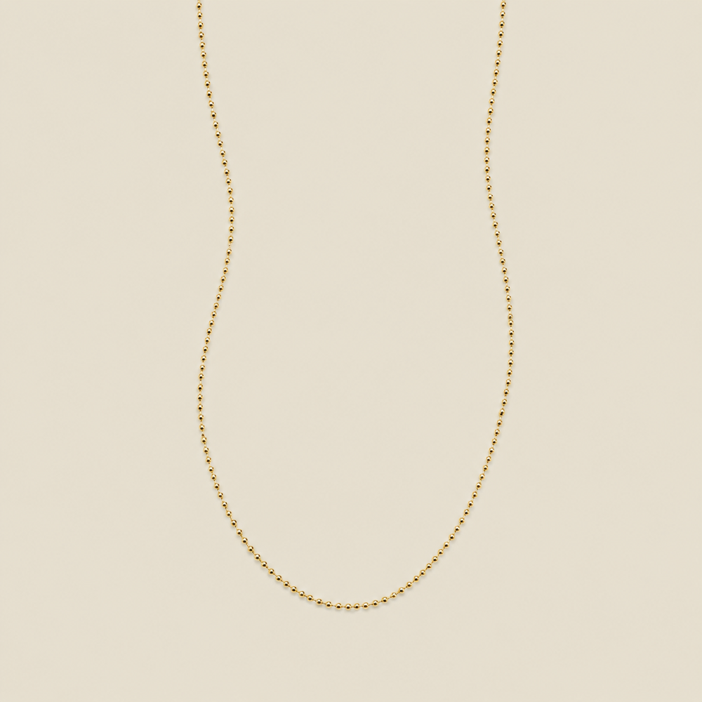 A delicate gold beaded chain necklace, suspended in a U-shape against a solid, warm beige background.