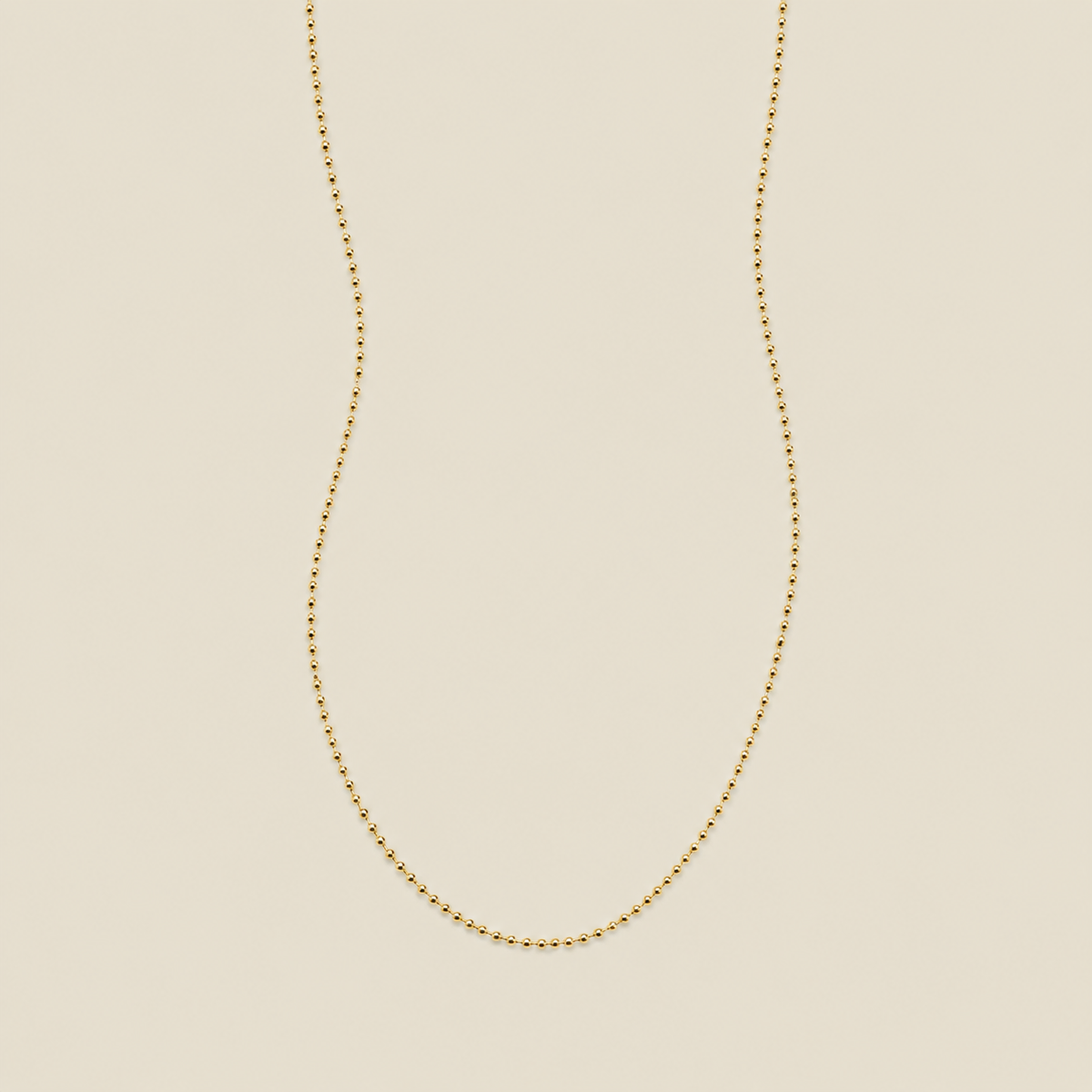 A delicate gold beaded chain necklace, suspended in a U-shape against a solid, warm beige background.