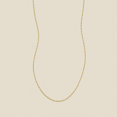 A delicate gold beaded chain necklace, suspended in a U-shape against a solid, warm beige background.
