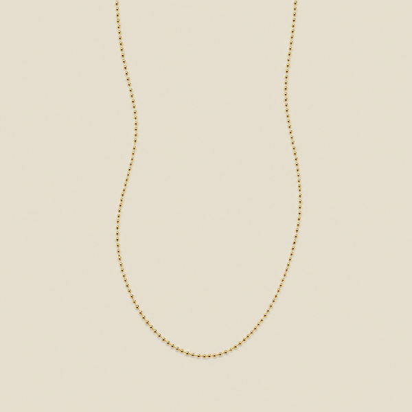 A delicate gold beaded chain necklace, suspended in a U-shape against a solid, warm beige background.