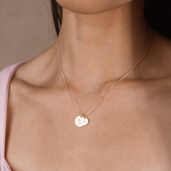 A close-up of a model's neck and chest wearing a delicate gold chain necklace with two small round engraved pendants, one of a hand drawn heart and the smaller disc of the letter 'K'.