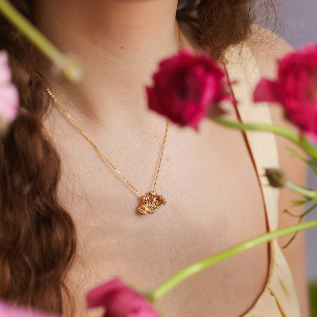 A close-up lifestyle image of a person wearing a gold pendant necklace with multiple gold flower charms, shot with multiple pink and red flowers blurred in the foreground and set against a purple background.