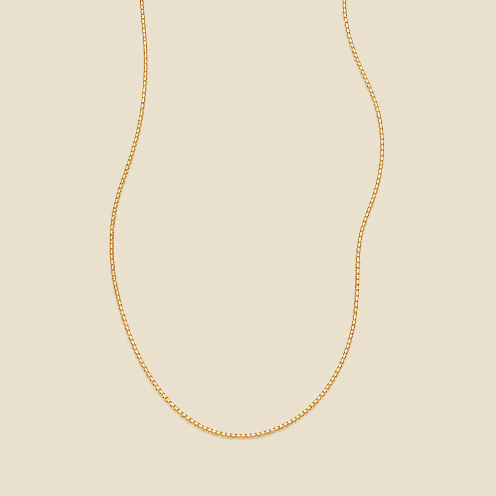 A delicate gold box chain necklace draped in a U-shape against a solid, warm beige background.