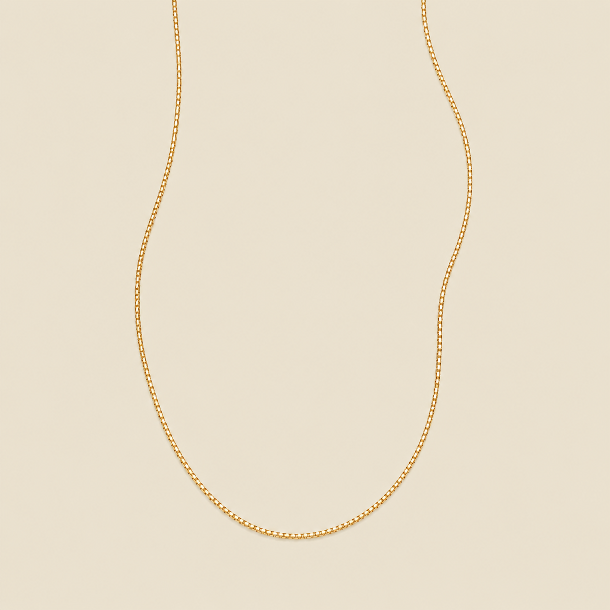 A delicate gold box chain necklace draped in a U-shape against a solid, warm beige background.