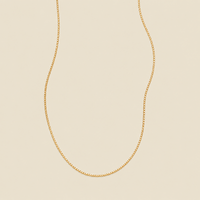 A delicate gold box chain necklace draped in a U-shape against a solid, warm beige background.