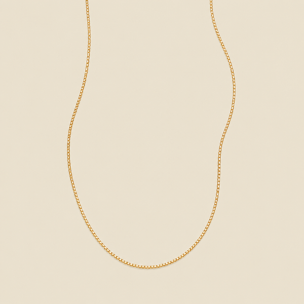A delicate gold box chain necklace draped in a U-shape against a solid, warm beige background.