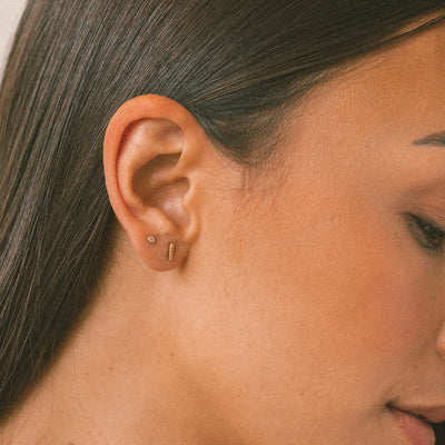 Close-up of a woman's ear wearing two small gold ear piercings, one bezel set clear cz stud and one short vertical bar, against her dark brown hair and warm skin tones.