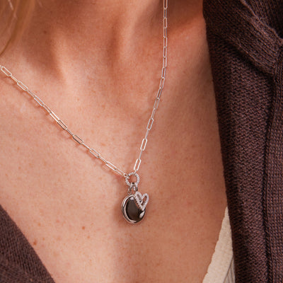A close-up of a person's neck and chest wearing a silver paperclip chain necklace with a silver oval locket pendant and a small silver heart charm with clear stones.