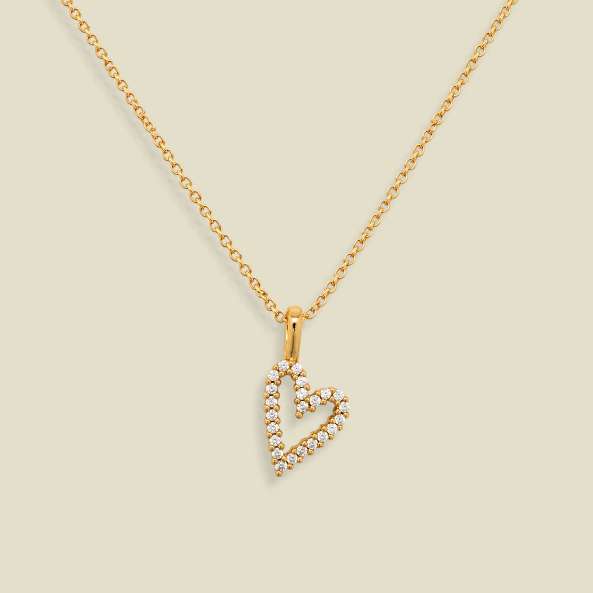 A gold chain necklace with a heart-shaped pendant outlined in small clear stones, displayed on a solid light beige background.