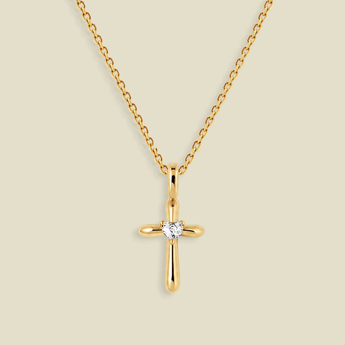 A gold cross pendant necklace with a small clear heart-shaped gemstone at the center, displayed on a light beige background.
