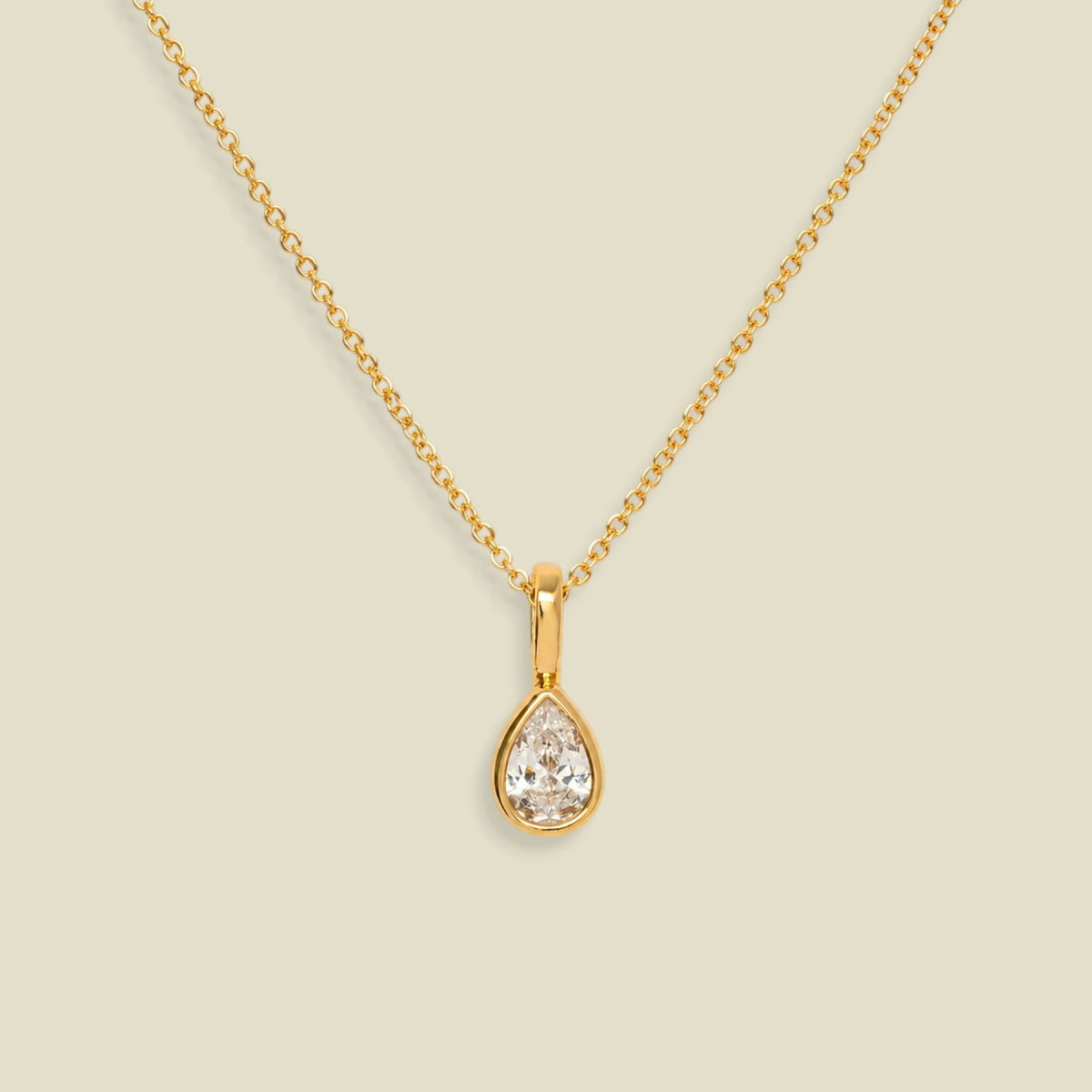 A gold chain necklace with a teardrop-shaped clear gemstone pendant in a gold bezel setting, displayed against a solid light beige background.