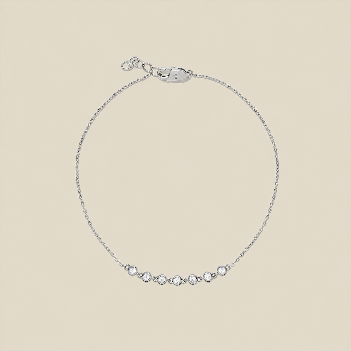 A delicate silver chain bracelet with a row of 7 small clear bezel-set round stones across the center, displayed on a solid light beige background.