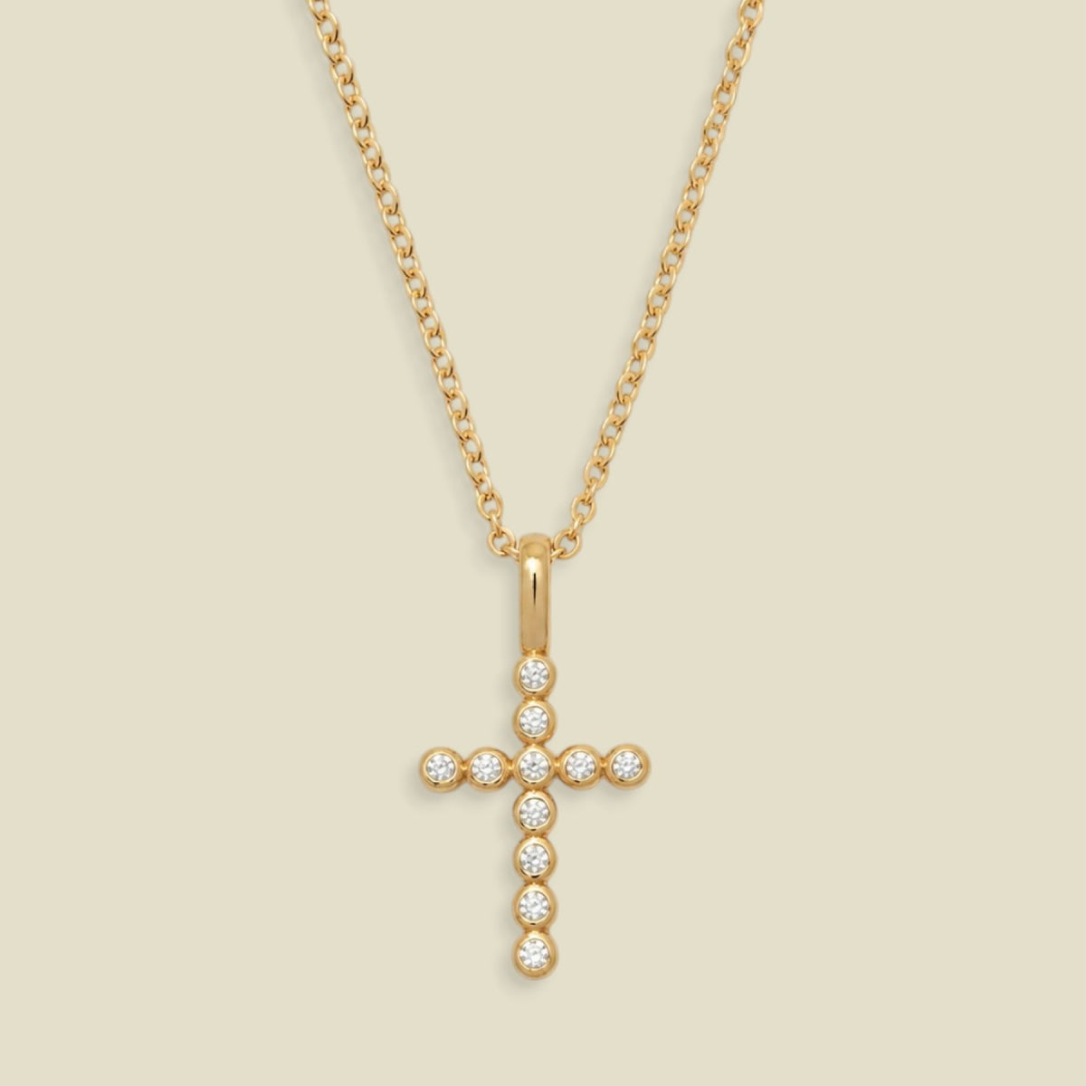 A gold cross pendant necklace adorned with small clear round bezel set gemstones, suspended from a delicate gold chain against a light beige background.