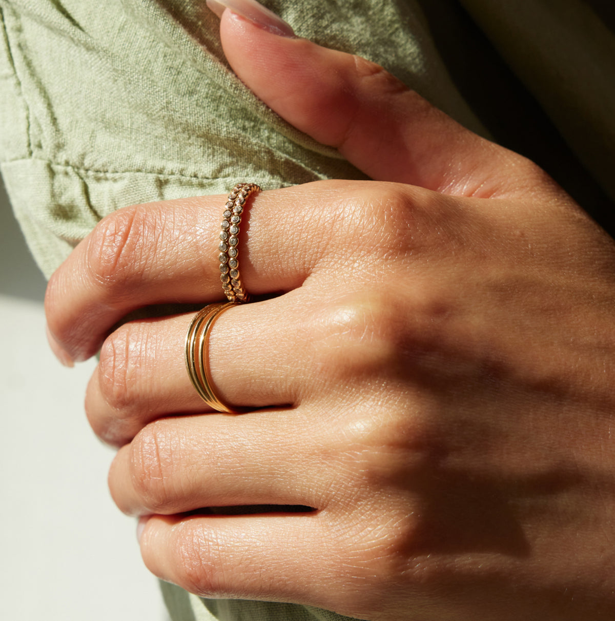 A close-up of a person's hand wearing four gold rings, including two textured beaded rings stacked on one another and two slim band rings stacked, against a soft olive green fabric background.