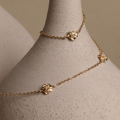 A lifestyle image of a dainty gold necklace with stationary textured flowers with small accent stones in the center along the chain, draped around a beige vase on a neutral background.