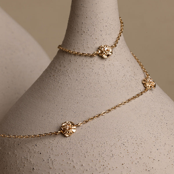 A lifestyle image of a dainty gold necklace with stationary textured flowers with small accent stones in the center along the chain, draped around a beige vase on a neutral background.
