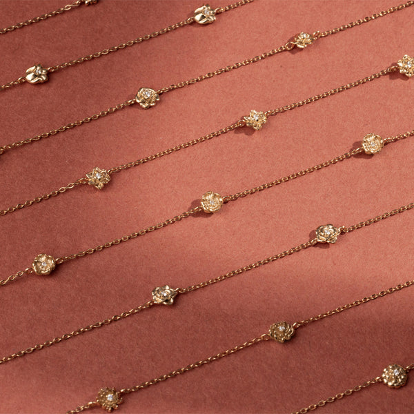 Eight delicate gold chain bracelets, each featuring ornate, textured gold flower charms with small clear stones, arranged side by side on a red background.