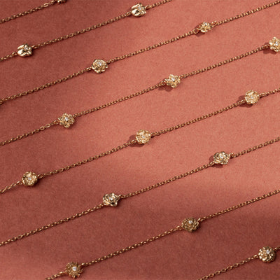 Eight delicate gold chain bracelets, each featuring ornate, textured gold flower charms with small clear stones, arranged side by side on a red background.