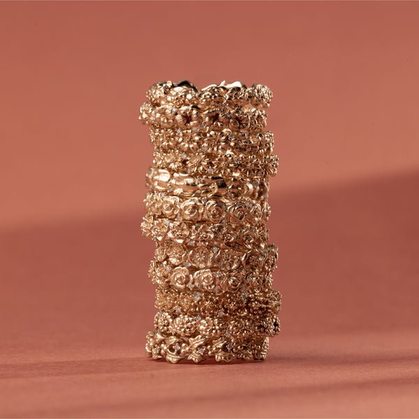 A vertical stack of 12 gold floral-etched ring bands with textured flowers and small clear gemstones, shown on a plain warm light red background. 