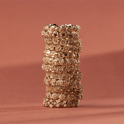 A vertical stack of 12 gold floral-etched ring bands with textured flowers and small clear gemstones, shown on a plain warm light red background. 