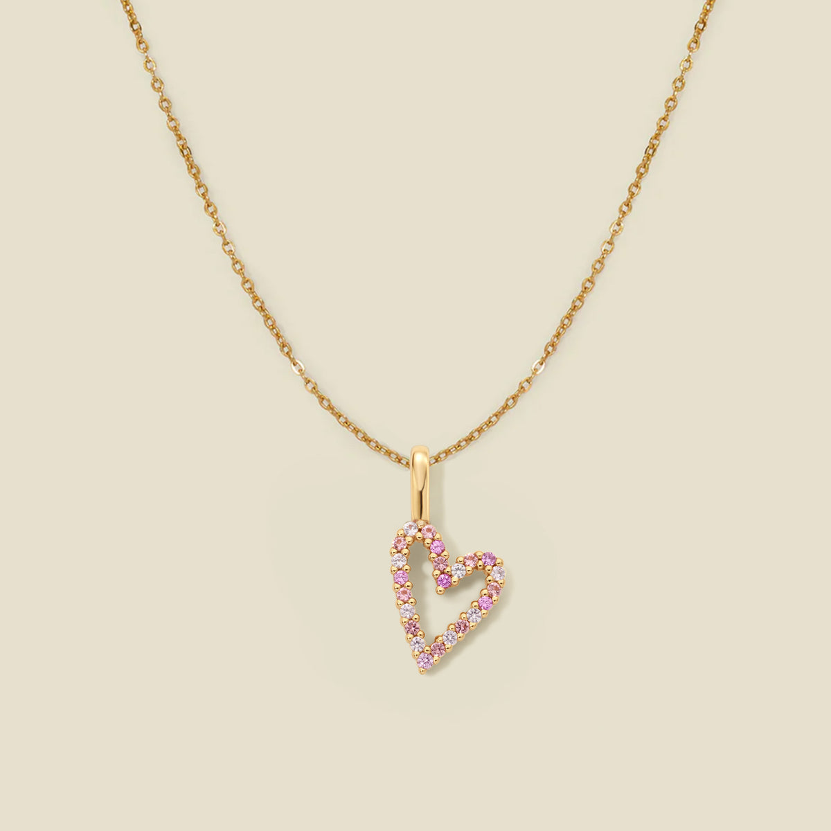 A delicate gold chain necklace featuring a heart-shaped pendant outlined with small pink gemstones, set against a solid light beige background.