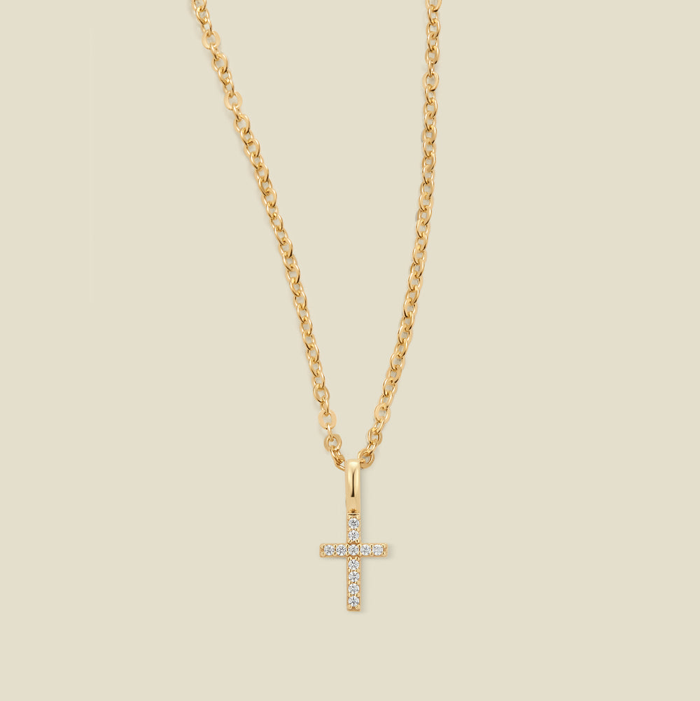 A gold cross pendant necklace with small clear stones set across the front,  suspended from a gold chain against a light beige background.