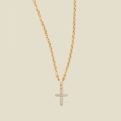 A gold cross pendant necklace with small clear stones set across the front,  suspended from a gold chain against a light beige background.