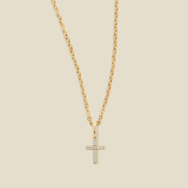 A gold cross pendant necklace with small clear stones set across the front,  suspended from a gold chain against a light beige background.