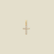 A small gold cross charm covered in small clear stones, set against a solid beige background.