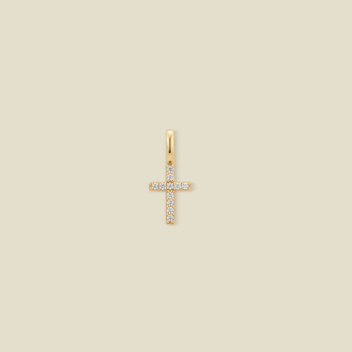A small gold cross charm covered in small clear stones, set against a solid beige background.