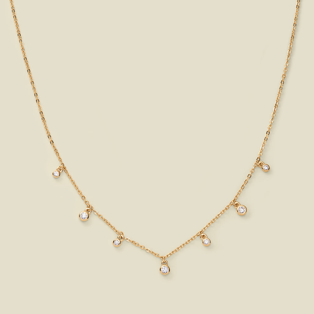 A dainty gold chain necklace with bezel-set clear, round gemstones stationed along it, set against a solid beige background.