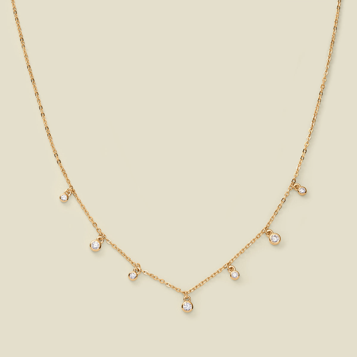 A dainty gold chain necklace with bezel-set clear, round gemstones stationed along it, set against a solid beige background.